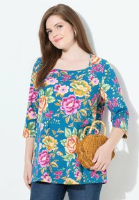 Blumige Tunika mit quadratischem Ausschnitt, lebendig blauer Hintergrund, ¾-Ärmel, mit rosa, gelben und grünen Blumen. Halte eine Korbtasche.