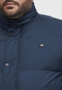 Navy blue padded jacket with a stand-up collar, featuring black buttons and a small black logo on the chest. Smooth fabric texture.