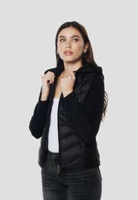 Črna puhasta jakna z kapuco, z našiljenim sprednjim delom in oprijetimi rokavi, iz gladkega blaga. Žepki z zadrgo na straneh.