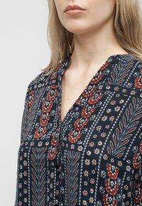 Γυναίκα με ανοιχτόξανθα μαλλιά που φοράει μπλε μπλούζα με σχέδια από κόκκινα, λευκά και μπλε λουλούδια και γεωμετρικά μοτίβα, εμφανιζόμενη από τους ώμους μέχρι το πηγούνι.