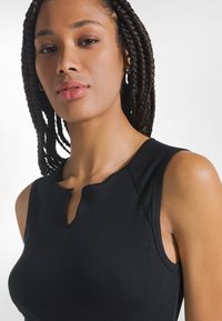 Mujer con el cabello trenzado, vestida con una blusa negra sin mangas con una pequeña abertura frontal, mirando confiada a la cámara sobre un fondo blanco.