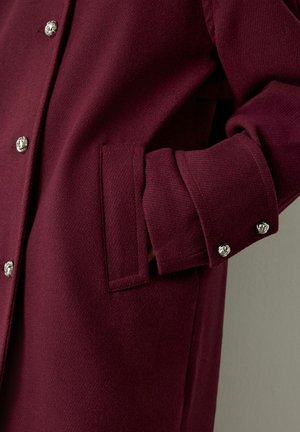 Close-up van een hand gedeeltelijk in de zijzak van een bordeauxrodejas met zilveren decoratieve knopen aan de voorkant en op de manchetten.