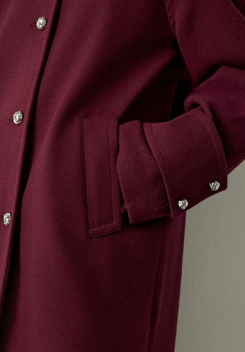 Close-up van een hand gedeeltelijk in de zijzak van een bordeauxrodejas met zilveren decoratieve knopen aan de voorkant en op de manchetten.