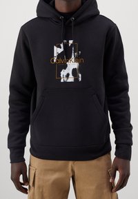 Felpa nera con cappuccio, tessuto strutturato, tasca frontale e logo Calvin Klein bianco e oro posizionato al centro del petto.
