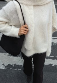Un pull doux et moelleux en tricot blanc superposé avec un sac noir, accompagné de pantalons sombres. La texture est douce, avec un design ample et des manches larges.