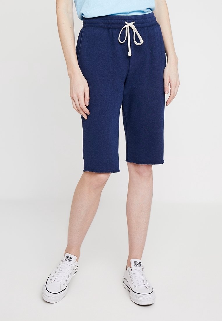 Person trägt marineblaue, knielange Shorts mit weißem Kordelzug und weiße Sneakers mit niedrigem Schaft und steht vor einem einfarbigen weißen Hintergrund.