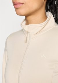 Veste beige à fermeture éclair avec un col montant. Texture du tissu lisse, avec un détail de logo subtil sur le côté gauche.