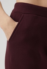 Burgunderfarbene Sweatpants mit geripptem Bund und Seitentasche. Weiche Textur mit glatter Oberfläche.