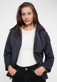 Marineblauwe hoodie gemaakt van soepel materiaal, met een lichte bontvoering, ritssluiting en elastische manchetten, gedragen over een witshirt.