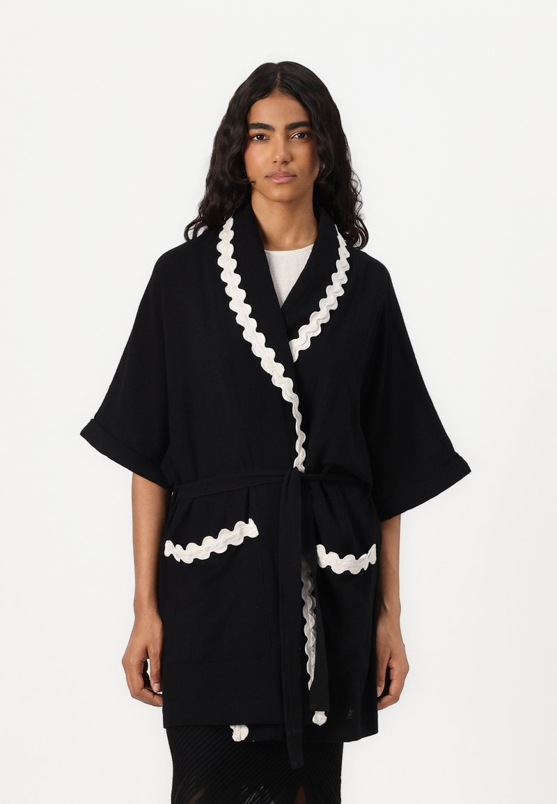 Robe noire à manches larges, avec une bordure en dentelle blanche le long du col et des poches, dotée d'une ceinture assortie pour la fermeture. Tissu texturé.