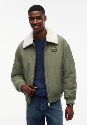 Homme souriant portant une veste bomber verte avec col en polaire blanche, pull bleu et jean sur fond blanc uni.