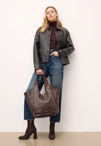 Rjava usnjena torba s dvojno ročajem, teksturirano površino in subtilnim zlatim logotipom. Uporablja se s temnimi kavbojkami in oprijetim zgornjim delom pod črnim jopičem.