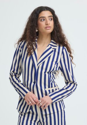 Kvinde med langt krøllet hår iført en blå og hvid lodretstribet blazer og matchende bukser, der kigger til siden med hænderne foldet.