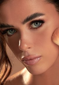 Primo piano di una donna con occhi azzurri e sopracciglia pronunciate che applica della cipria sulla guancia con un pennello.