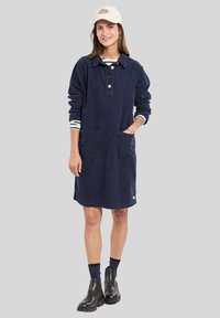 Robe en corduroy marine avec de longues manches, col boutonné, deux poches avant et revers rayés ; associée à des bottines noires et une casquette.