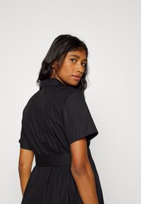 Vestido preto de camisa com mangas curtas, colarinho e laço nas costas. Feito de tecido suave com cintura ajustada e saia rodada. Design simples e elegante.