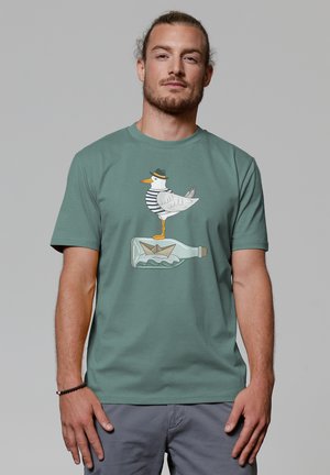 Groene katoenen T-shirt met een cartoon-eend in een gestreept shirt, staande op een fles met een papieren bootje erin, op een effen achtergrond.