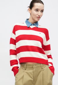 Πλεκτό πουλόβερ με κόκκινες και λευκές ρίγες και ριμπ λαιμόκοψη, φορεμένο πάνω από ελαφρύ πουκάμισο από τζιν με γιακά. Μπεζ παντελόνι με πλαϊνές τσέπες.