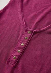 Canotta aderente in magenta profondo, con scollo a V e inserto con cinque bottoni in ottone. Texture a maglia con finitura liscia.