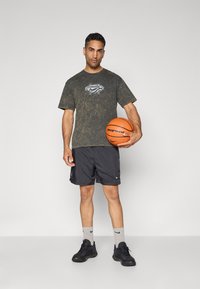 Mørkegrøn tie-dye t-shirt med et centralt Nike-logo, parret med sorte shorts, grå sokker og sorte sneakers, mens han holder en orange basketball.