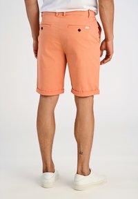 Shorts de couleur pêche avec des revers, en tissu léger. Deux poches arrière avec des boutons noirs et une petite étiquette de marque.