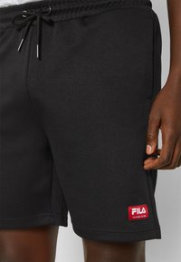 Shorts sportivi neri realizzati in tessuto testurizzato, dotati di una vita elasticizzata con coulisse e una patch rossa con logo sul lato sinistro inferiore.