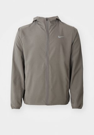 Grå vindjakke med en tætsiddende hætte, lynlås fortil og elastiske manchetter. Har et subtilt Nike-logo på brystet. Glat, mat tekstur.