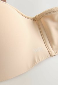 Beige bh van glad materiaal, met onderdraadondersteuning en verstelbare banden. Heeft een subtiele logo op de zijkant. Geëxposeerde stikseldetails.