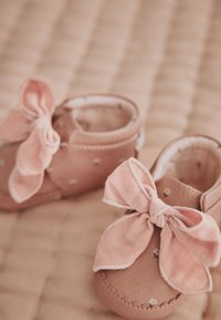 Des chaussons de bébé roses doux avec de grands nœuds en tissu reposant sur une surface beige matelassée.