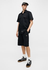 Jeune homme portant une chemise noire à manches courtes avec boutons, un short cargo noir, une casquette noire, des chaussettes blanches et des baskets noires à semelles en gomme, debout devant un fond uni.
