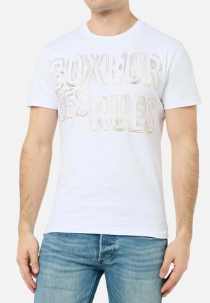 Mann trägt weißes kurzärmeliges T-Shirt mit dem Text "BOXEUR DES RUES" in großen Umrissbuchstaben und blauer Jeans, steht mit entspannten Armen.