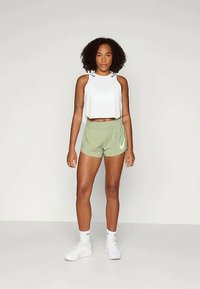 Γυναίκα με σγουρά μαλλιά που φοράει λευκό χωρίς μανίκια κορμάκι, ανοιχτά πράσινα αθλητικά σορτς, λευκές κάλτσες και λευκά αθλητικά παπούτσια, στέκεται μπροστά από απλό φόντο.