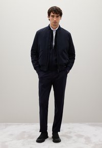 Giacca bomber navy con zip e tasche laterali sopra un maglione con zip abbinato, abbinata a pantaloni navy slim-fit e scarpe slip-on nere.