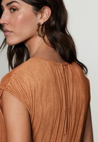 Haut texturé de couleur rouille avec un motif à rayures subtile, présentant des manches courtes et une fermeture à bouton au niveau du col au dos.