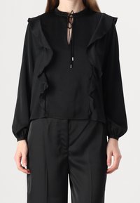 Blouse noire à col haut, ornée de volants sur le devant et les manches, confectionnée dans un tissu lisse, associée à un pantalon assorti.