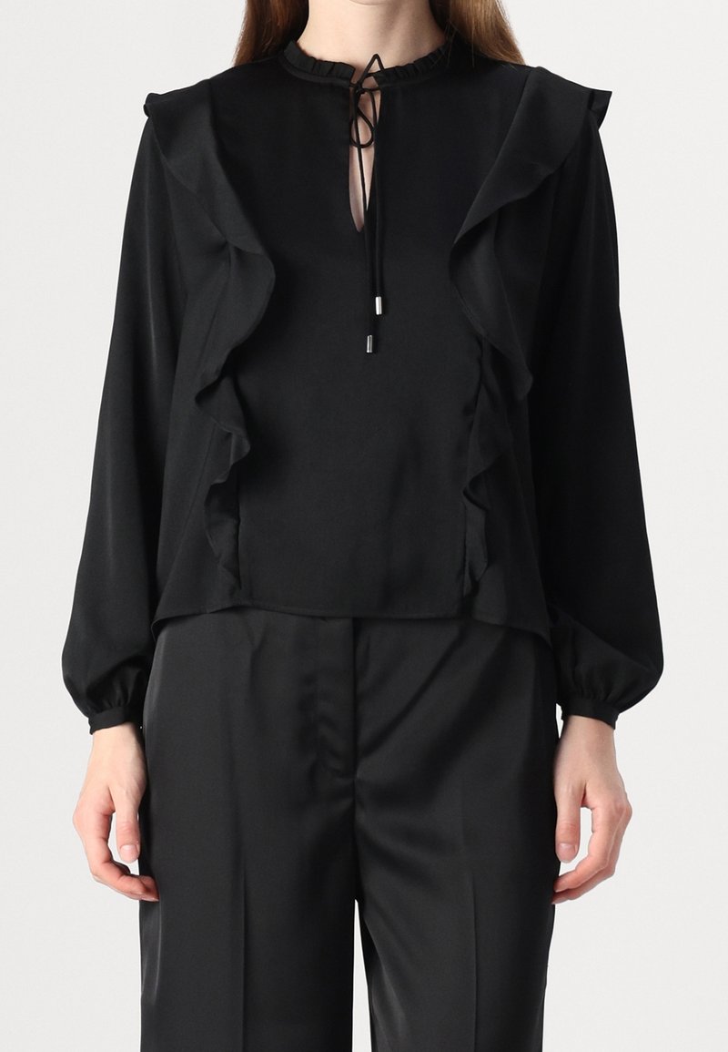 Blouse noire à col haut, ornée de volants sur le devant et les manches, confectionnée dans un tissu lisse, associée à un pantalon assorti.