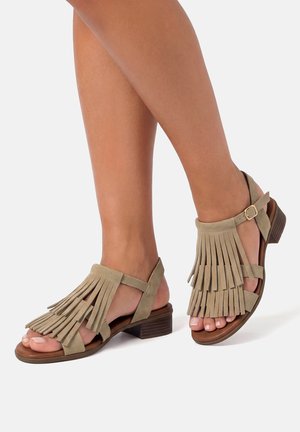 Sandalias de ante color cuero con flecos en capas, correa en el tobillo y tacón bajo apilado, usadas en pies cruzados sobre fondo blanco.