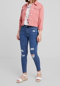 Donna che indossa una giacca di jeans rosa, una blusa floreale, jeans skinny blu strappati e sandali con tacco a cuneo bianchi, in piedi su uno sfondo semplice.