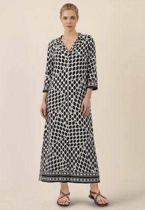 Femme portant une longue robe à motifs noir et blanc avec des manches trois-quarts et des sandales marron, debout devant un fond uni.