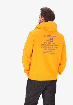 Uomo di spalle che indossa una felpa con cappuccio arancione brillante con testo tecnico stampato sul retro e pantaloni scuri su sfondo bianco.