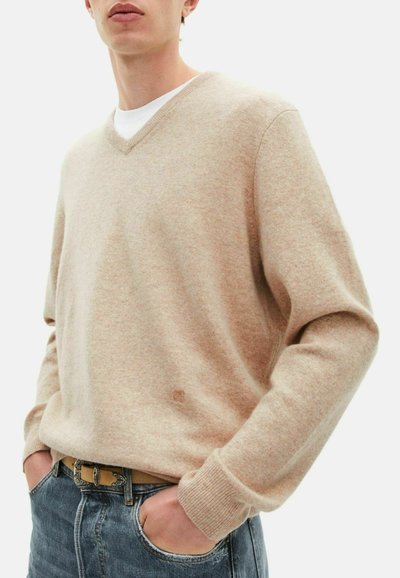 Suéter de punto beige con cuello en V, mangas largas y puños acanalados. Lleva puesto sobre una camiseta blanca, combinado con jeans de mezclilla azules.
