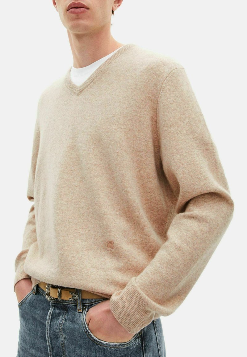 Beige Strickpullover mit V-Ausschnitt, langen Ärmeln und gerippten Bündchen. Getragen über einem weißen T-Shirt, kombiniert mit blauen Jeans.