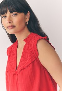 Rote Bluse mit bestickten Details, gerafften Schulternähten und lockerem Sitz. Hergestellt aus leichtem Stoff mit strukturierter Oberfläche.