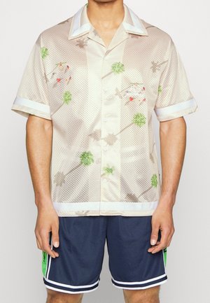 Kurzärmliges, beiges Mesh-Shirt mit weißem Kragen und Bündchen, das mit grünen und mehrfarbigen Blumenmustern verziert ist. Getragen mit marineblauen Shorts.