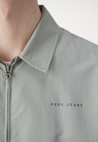Jachetă verde deschis cu închidere cu fermoar și guler ascuțit; are un finisaj mat și branding 'PEPE JEANS' pe partea stângă.