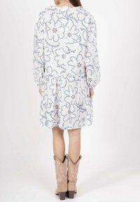 Robe blanche avec des motifs floraux bleus, manches longues, ourlet froncé et fermeture à boutons au dos ; associée à des bottes de cowboy beiges.