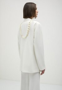 Blazer bianco su misura con vestibilità rilassata, abbinato a pantaloni coordinati. Presenta una collana di perle drappeggiata sulle spalle.