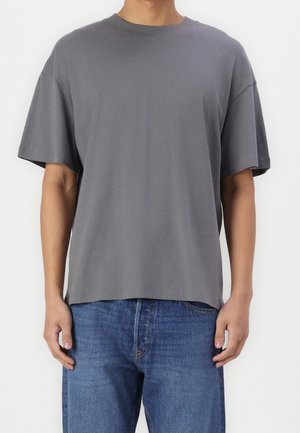 Person iført en løstsiddende grå t-shirt med korte ærmer og blå jeans, stående mod en ensfarvet hvid baggrund.