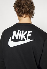 Felpa nera con un grande logo bianco "NIKE" e un baffo stampati sul retro. Collo a giro e maniche lunghe; texture del tessuto morbida.