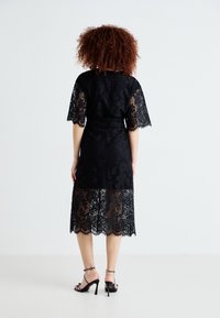 Robe en dentelle noire à manches courtes et ourlet festonné. Présente une couche intérieure en slip, un revêtement transparent, et des talons hauts avec des accents cloutés.